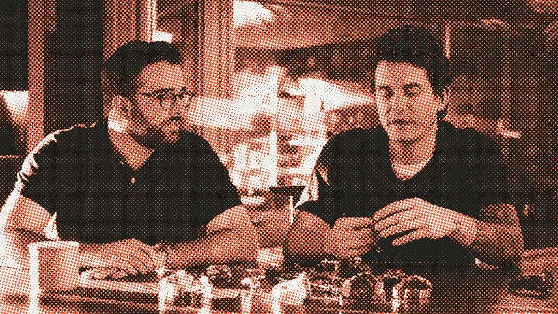 Two men at a wooden table covered in watches, one of them handling a watch while the other looks on